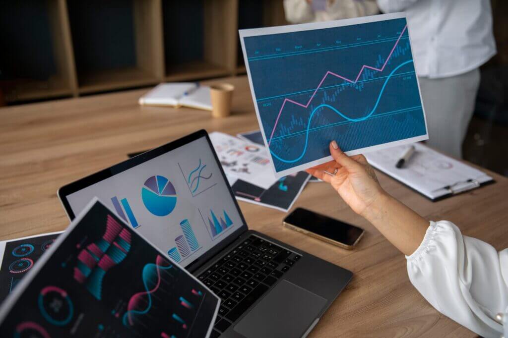 A person’s hand holding up a printed graph with trend lines in front of a laptop displaying various charts and data analysis, symbolizing ongoing model refinement and support for sustained accuracy in a business or analytics setting