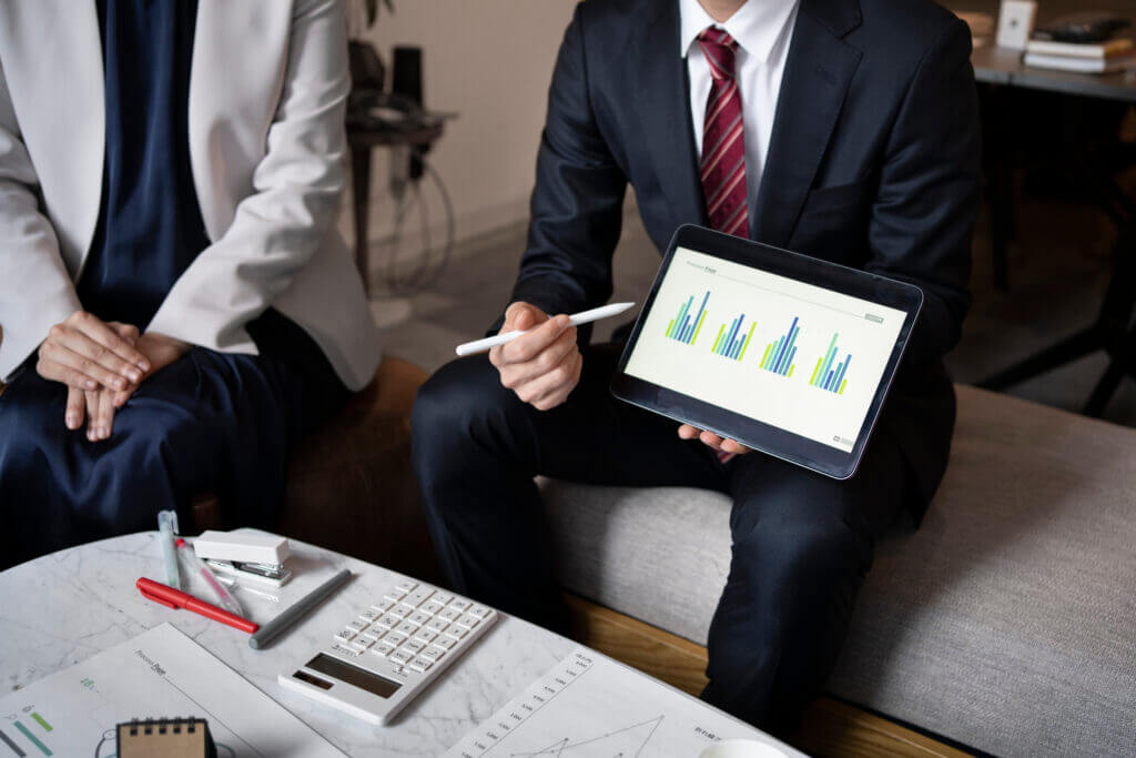 A professional meeting with one individual holding a tablet showing bar graphs indicative of financial analytics, alongside various business analysis tools on the table.