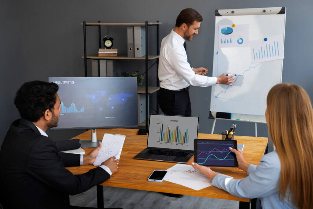 Healthcare professionals collaborate around a conference table, analyzing financial data on laptops and monitors while one presents revenue forecasts on a flip chart, highlighting the strategic use of predictive models in healthcare finance