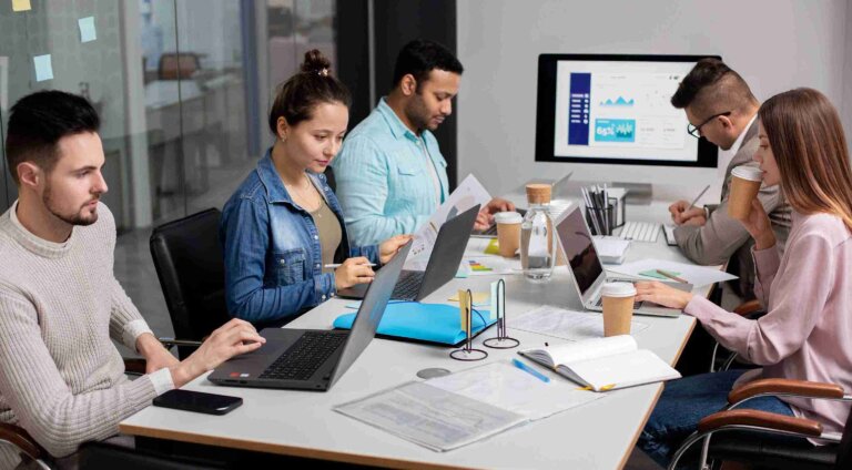 A group of professionals engaged in medical coding and auditing tasks at a conference table with laptops, documents, and a presentation screen in the background.